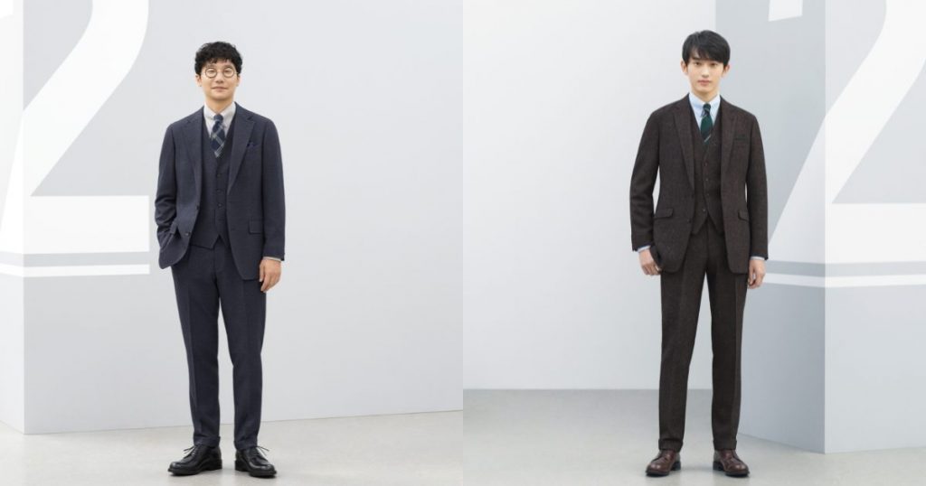 杉野さん、前原さんの２人が、スリーピーススーツスタイルで立っている。向かって左側の前原さんは、ネイビー系のスリーピーススーツ。ブラウン系（ベージュ）シャツに、同じくネイビー系の大振りのチェックのネクタイを合わせている。ブラックシューズ。
向かって右側の杉野さんは、ブラウン系のスリーピーススーツスタイル。淡いブルー系のシャツに、グリーン系の大きなストライプ柄ネクタイを合わせている。シューズもブラウン。
背景は淡いグレー。
壁面に数字の”2”の柄が施されている。
都会的なイメージの画像。